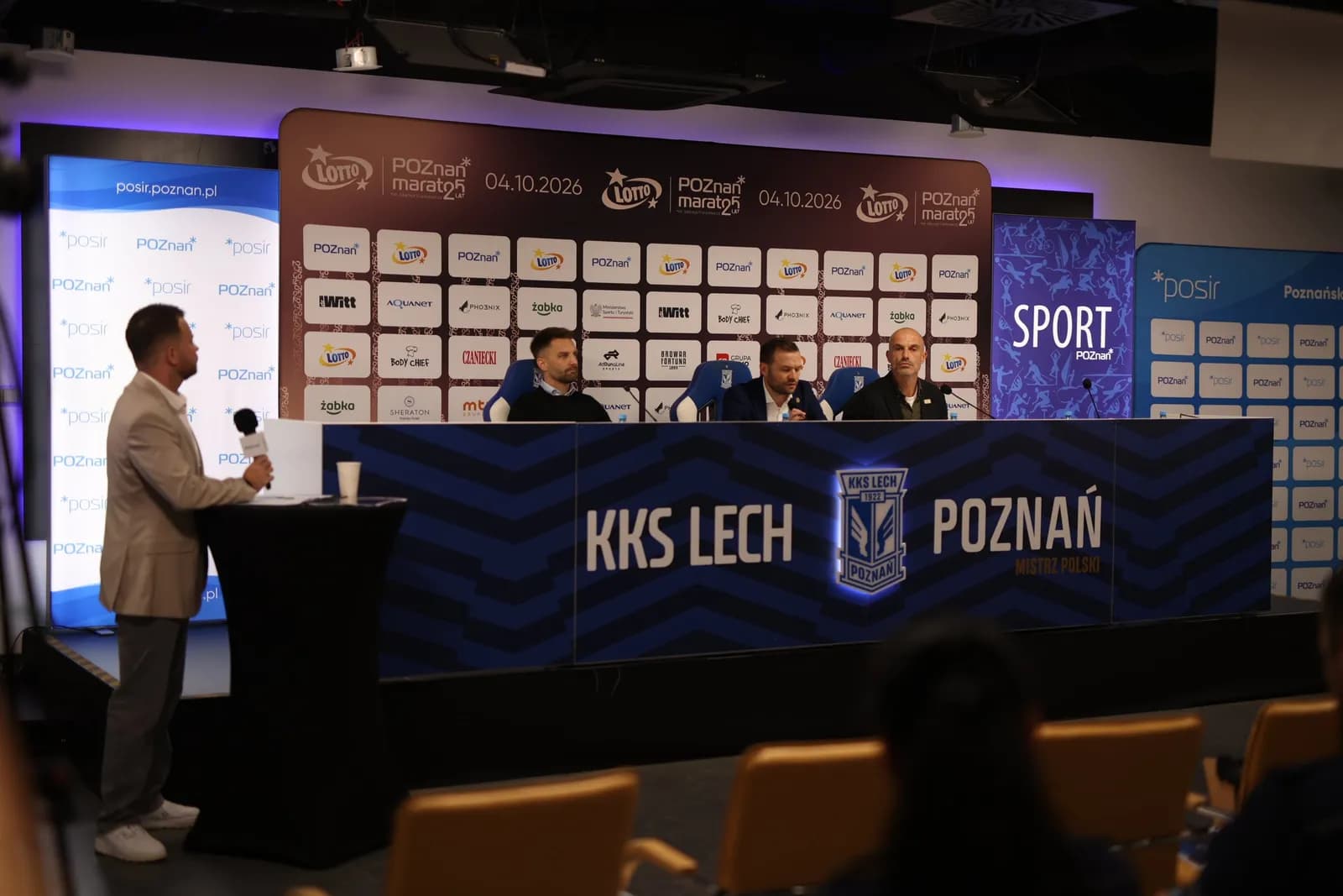 A man in a beige suit stands at a podium speaking into a microphone, facing three men seated at a long table behind him, with event branding and sponsor logos on the wall.
