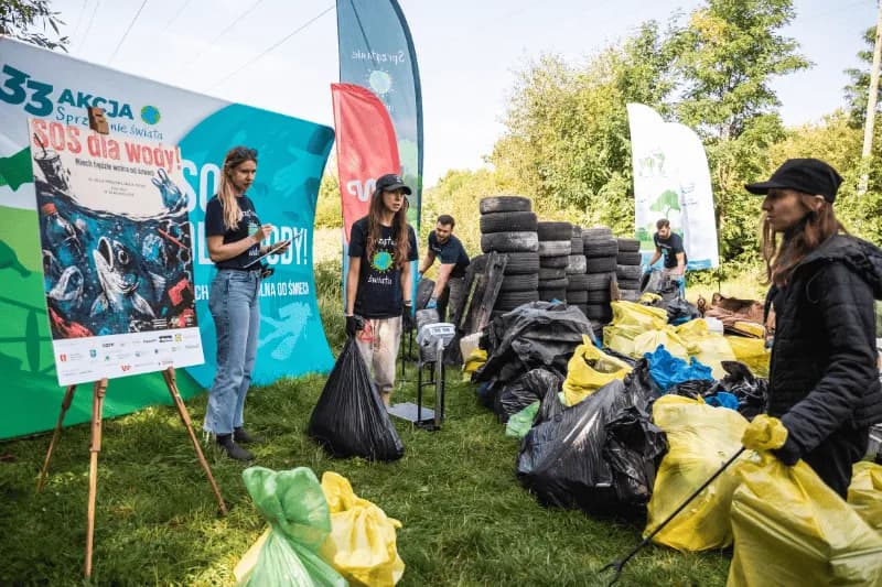 Volunteers are collecting trash, sorting it into different colored bags, with a large poster promoting water conservation visible in the background.