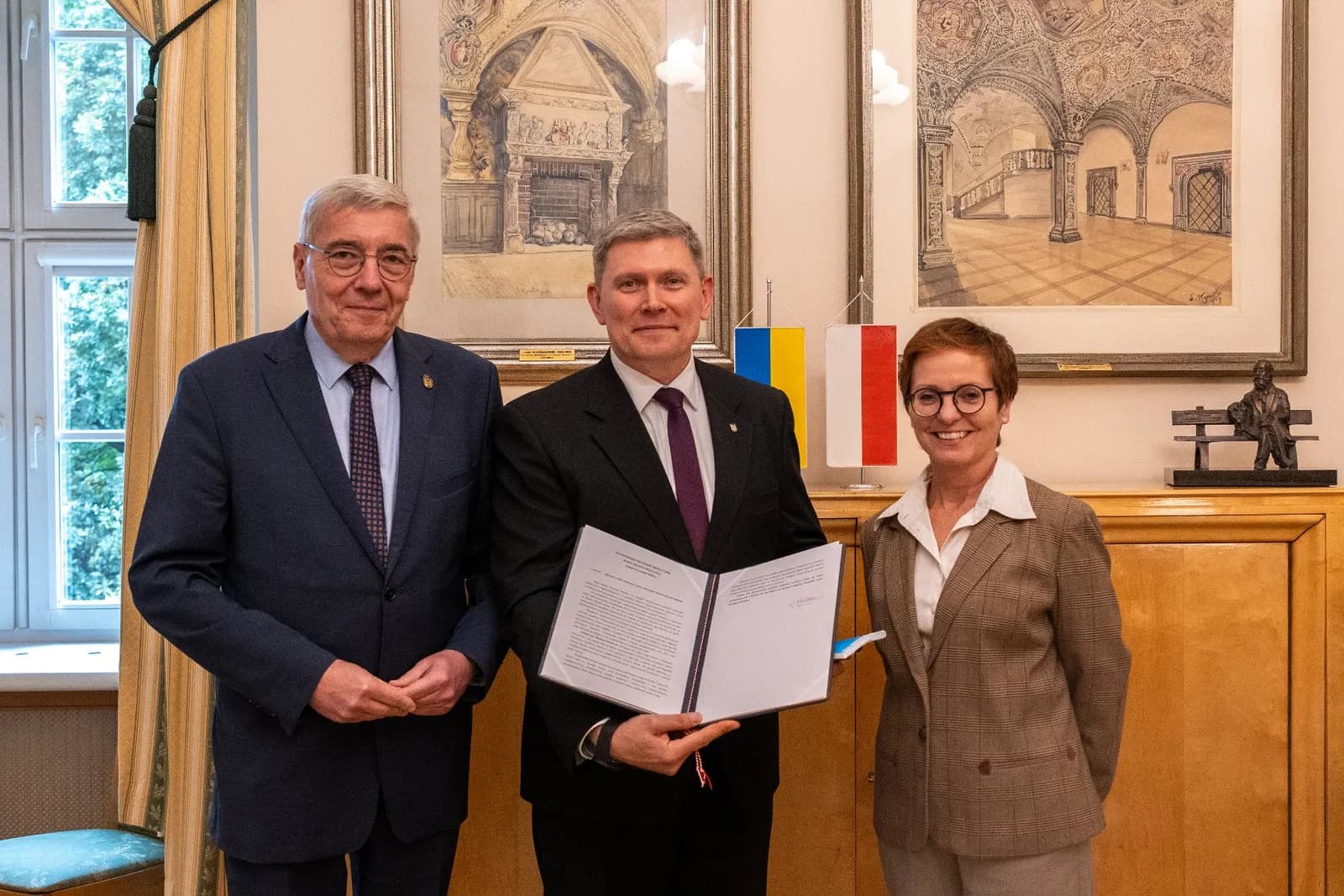 Three people, two men and one woman, in business attire standing in a formal room, with the man in the middle holding an open document.
